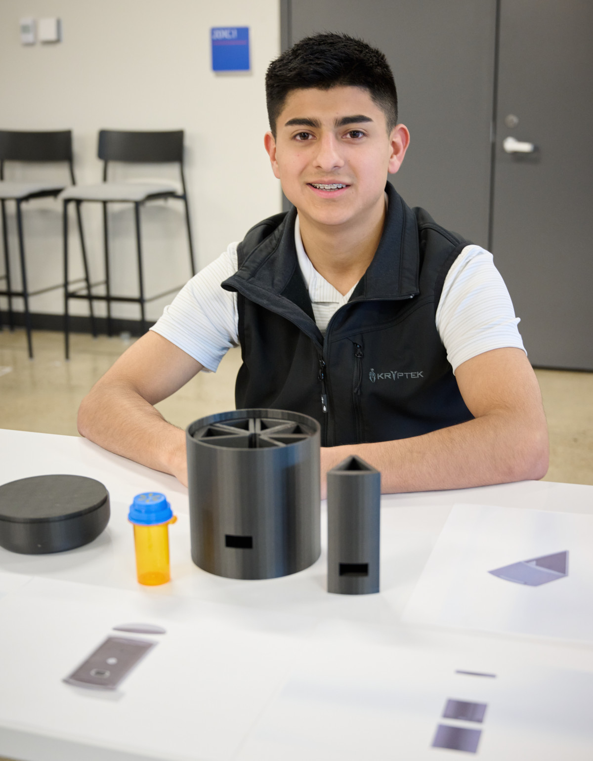Man in white shirt and black vest sitting at table with medicine container parts in front of him.